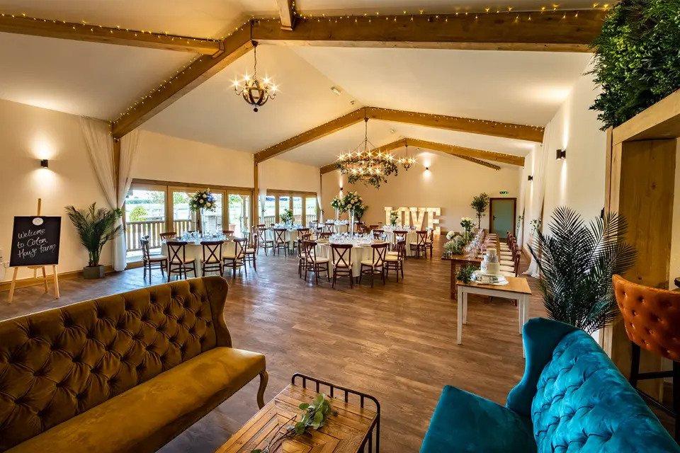 Room set for wedding meal with plush velvet sofas, wooden floors, chandeliers, white round tables, wooden chairs, fairy lights, floral centrepieces and LED letters spelling LOVE