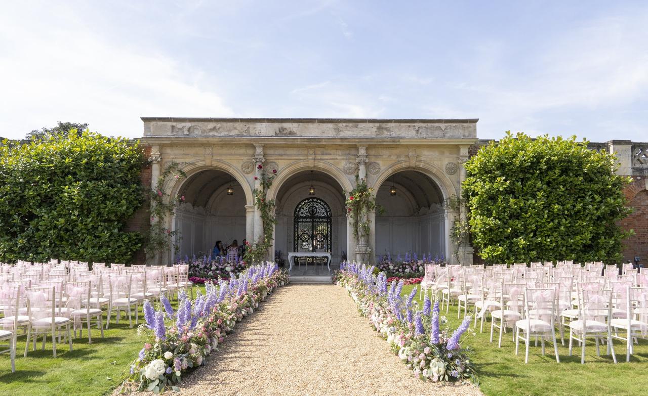 An outdoor ceremony set-up with floral-lined aisle and stone arches.