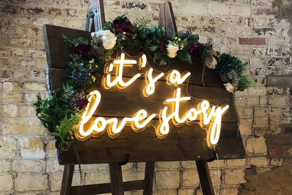A dark wooden wedding sign with green foliage and neon letters that say 