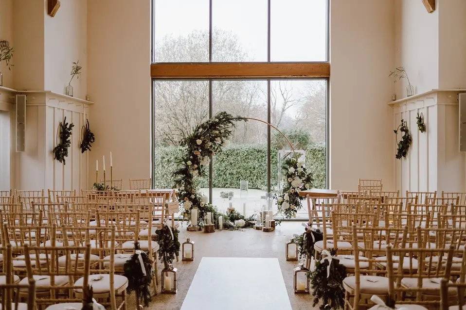 Room set for wedding ceremony with wooden chairs, pillar candles in lantern boxes, foliage decor, floor-to-ceiling windows and a floral hoop