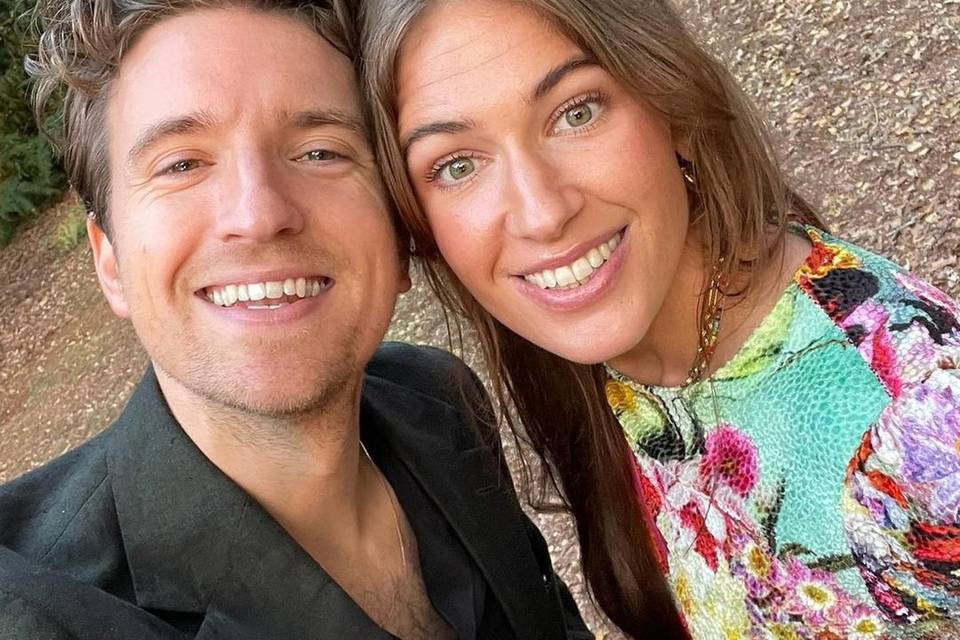 Greg James and Bella Mackie take a selfie. He is wearing a smart black jacket and she is wearing a colourful dress