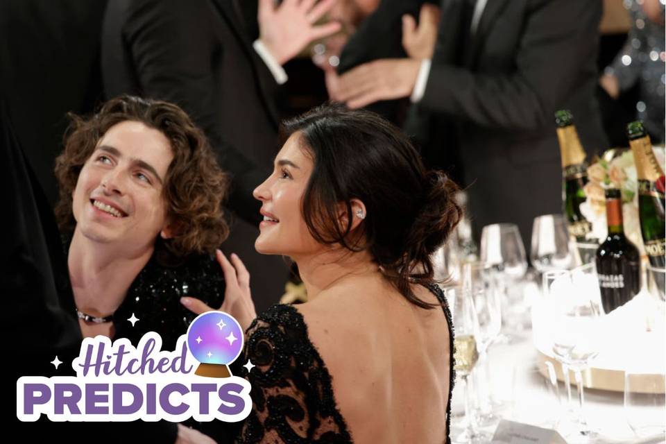 Kylie Jenner wearing a black, low back lace dress sitting at a table for the Golden Globes smiling to the left, placing one hand on Timothee Chalamet's shoulder who is also smiling and wearing a black, sparkly suit