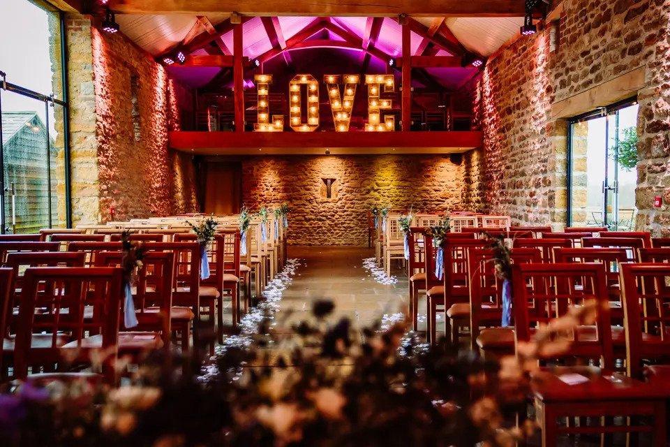 Rustic barn room set up for wedding ceremony with wooden chairs, wooden beams, exposed brick and wooden chairs with an aisle lined with flower petals