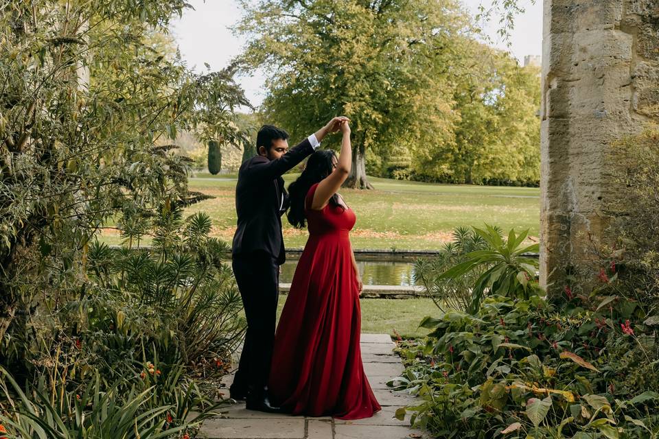 A couple in black suit and red dress dance in front of a backdrop of greenery and a lake