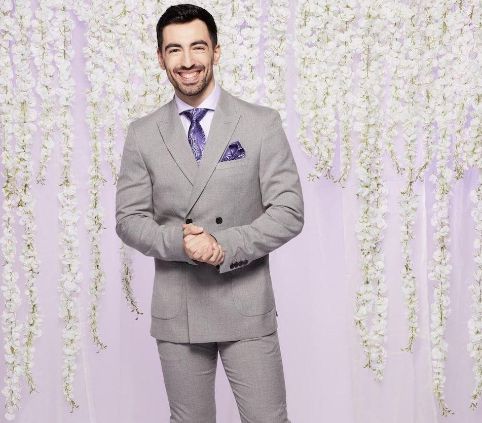 mafs uk groom thomas wears a grey suit and purple tie in front of a lilac backdrop with white hanging flowers