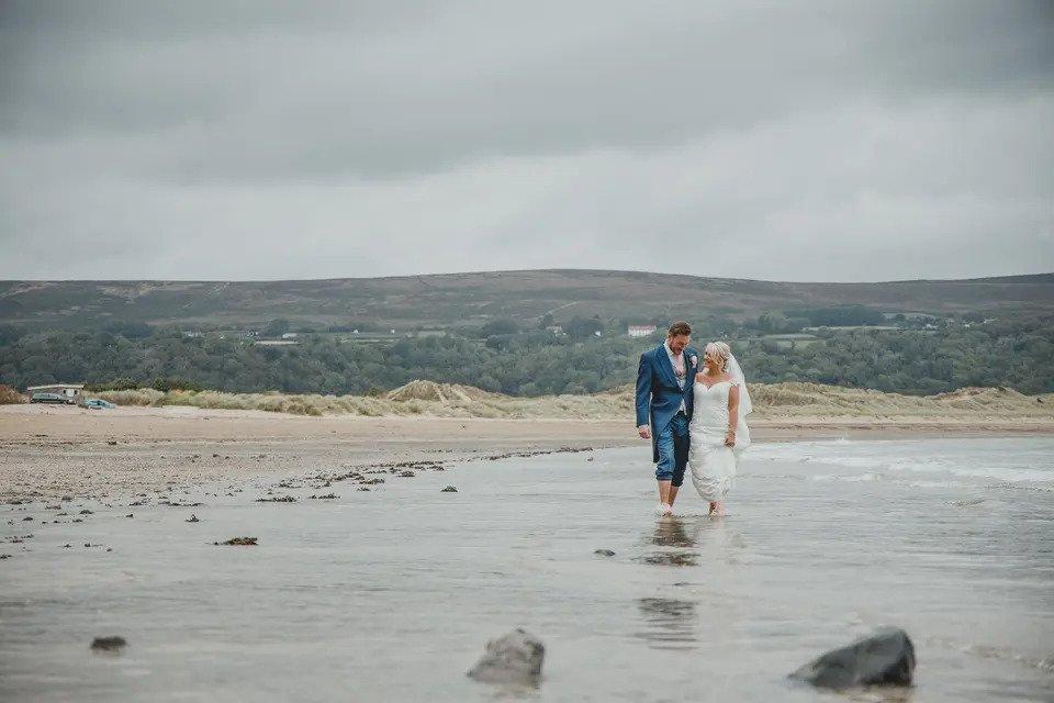Bride and groom photographed from a distance walking hand in hand across a beach in Wales