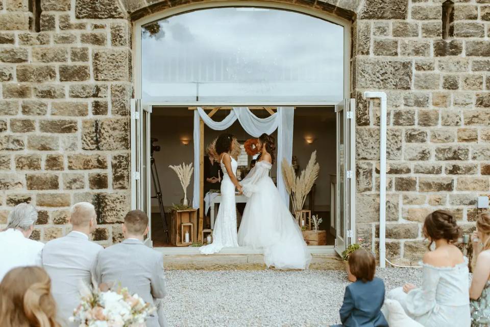 two brides hold hands during their wedding ceremony which is outside a rustic brick barn