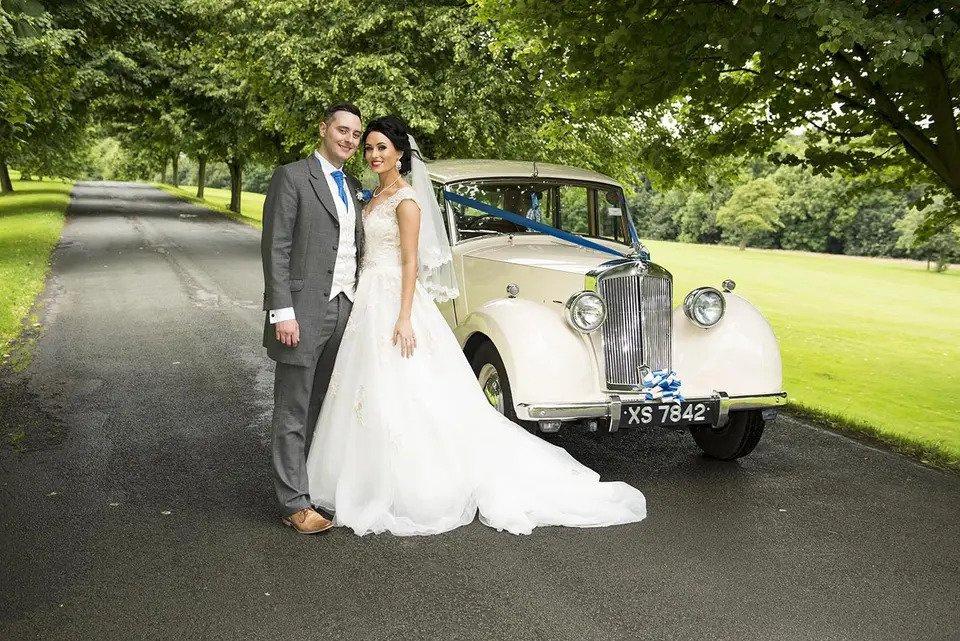 Bride and groom smile at the camera on a verdant driveway with a classic car
