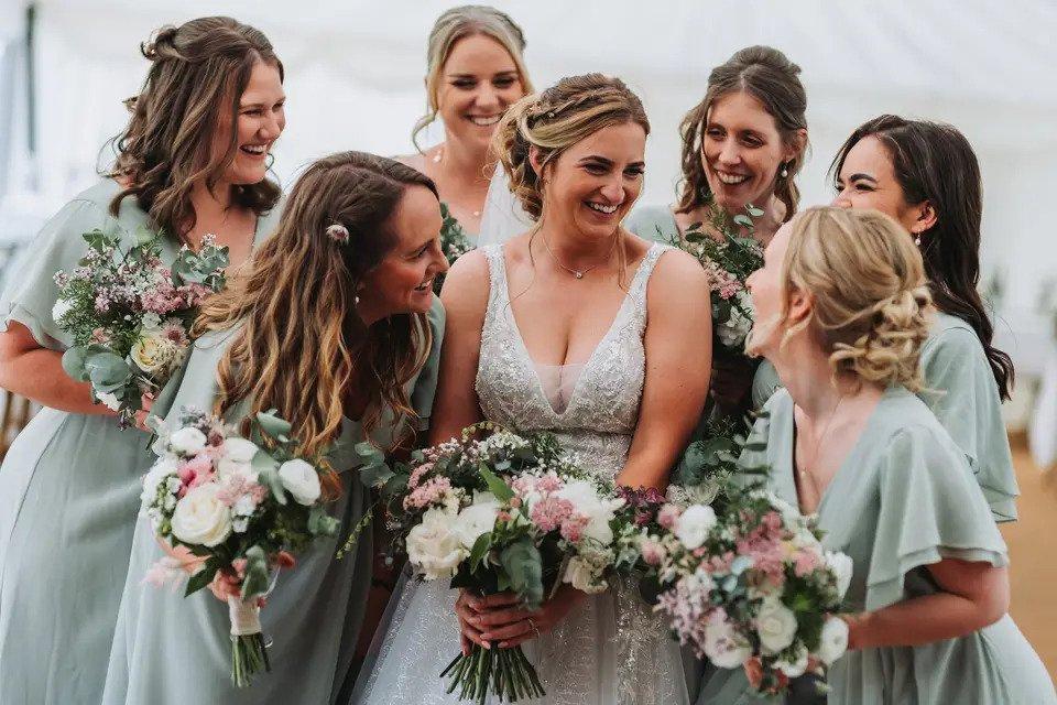 Bride laughing as she's closely surrounded six bridesmaids in pastel green bridesmaid dresses