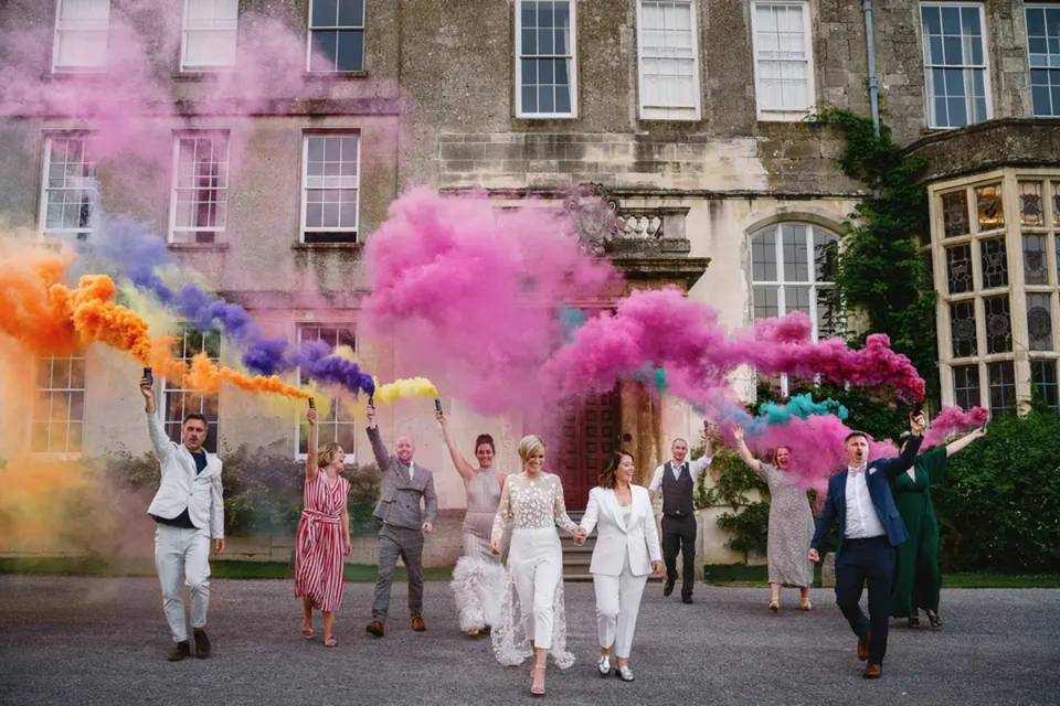 Two brides walking out of their wedding venue hand in hand with their guests behind them letting off colourful smoke bombs 