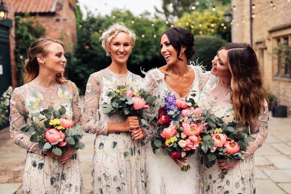 A bride and her bridesmaids wearing beaded long sleeved dresses holding gorgeous floral bouquets at a brick wedding venue
