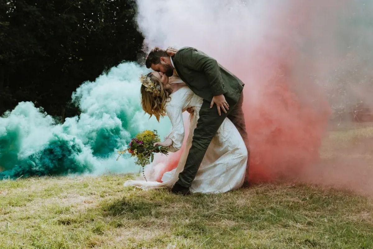 a groom holding his bride's back as she leans very far backwards, surrounded by teal and pink smoke
