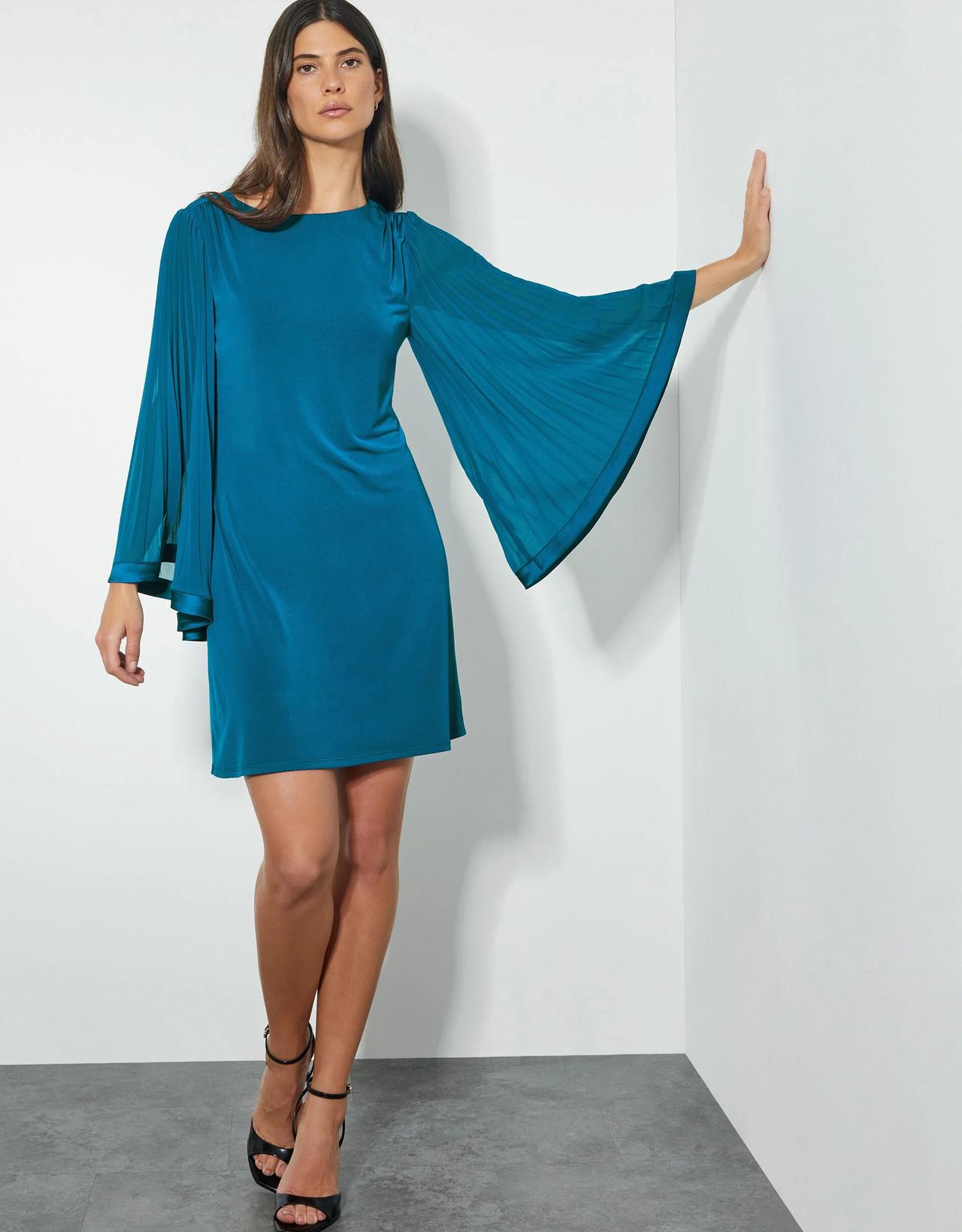 A wedding guest wearing a teal-coloured mini dress with dramatic long cape sleeves.