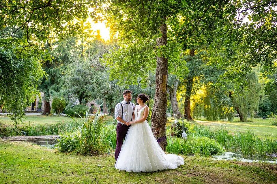 Bride and groom holding hands in the grounds of a wedding venue, posing looking at each other in front of a tree