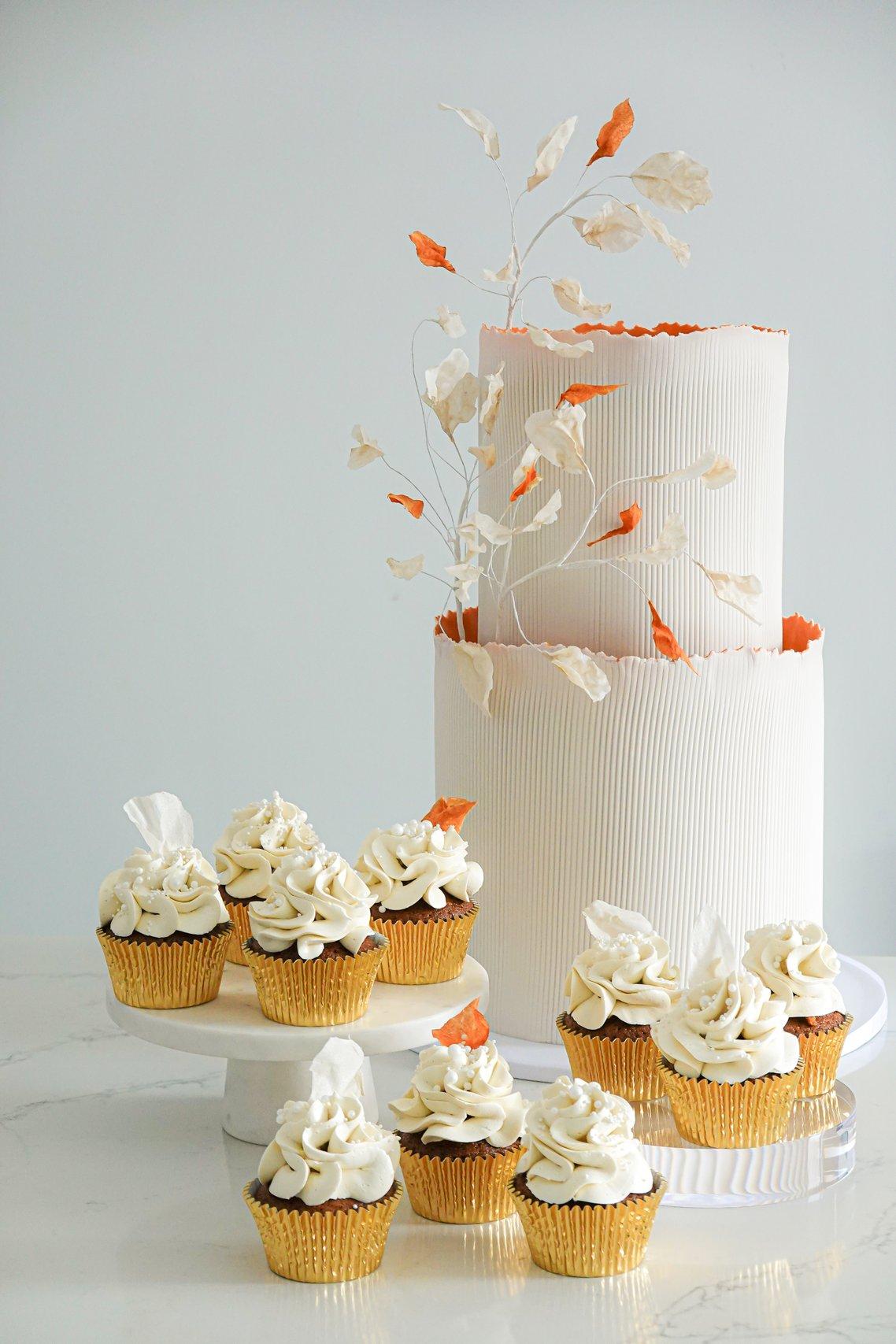 A two-tier white iced wedding cake with vertical line detail surrounded by some simple cupcakes in gold cases.