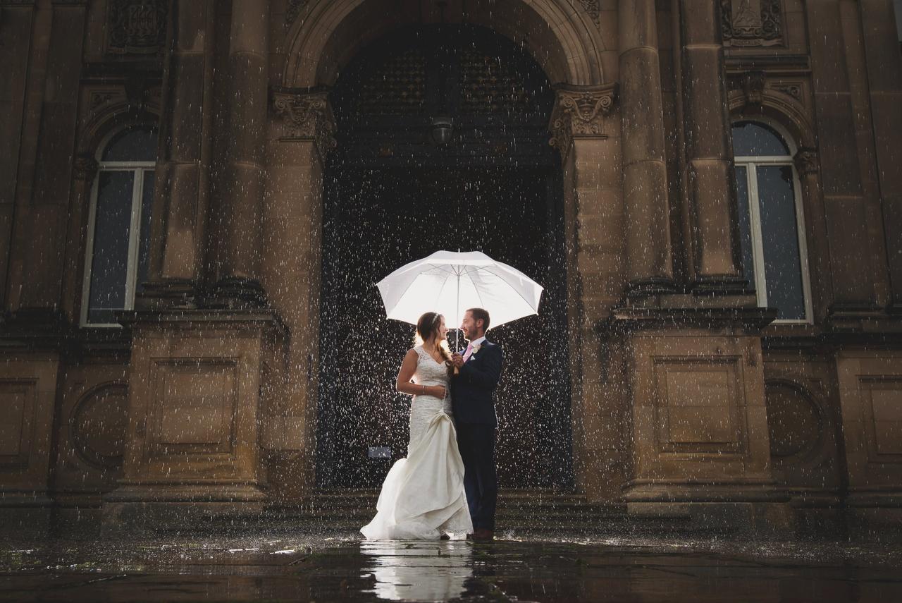 A couple standing outside the beautiful stone wedding venue with an umbrella in the rain