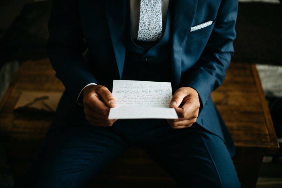 A man wearing a wedding suit visible from the shoulders down holding a wedding speech and reading over it in order to practise his wedding speech introduction