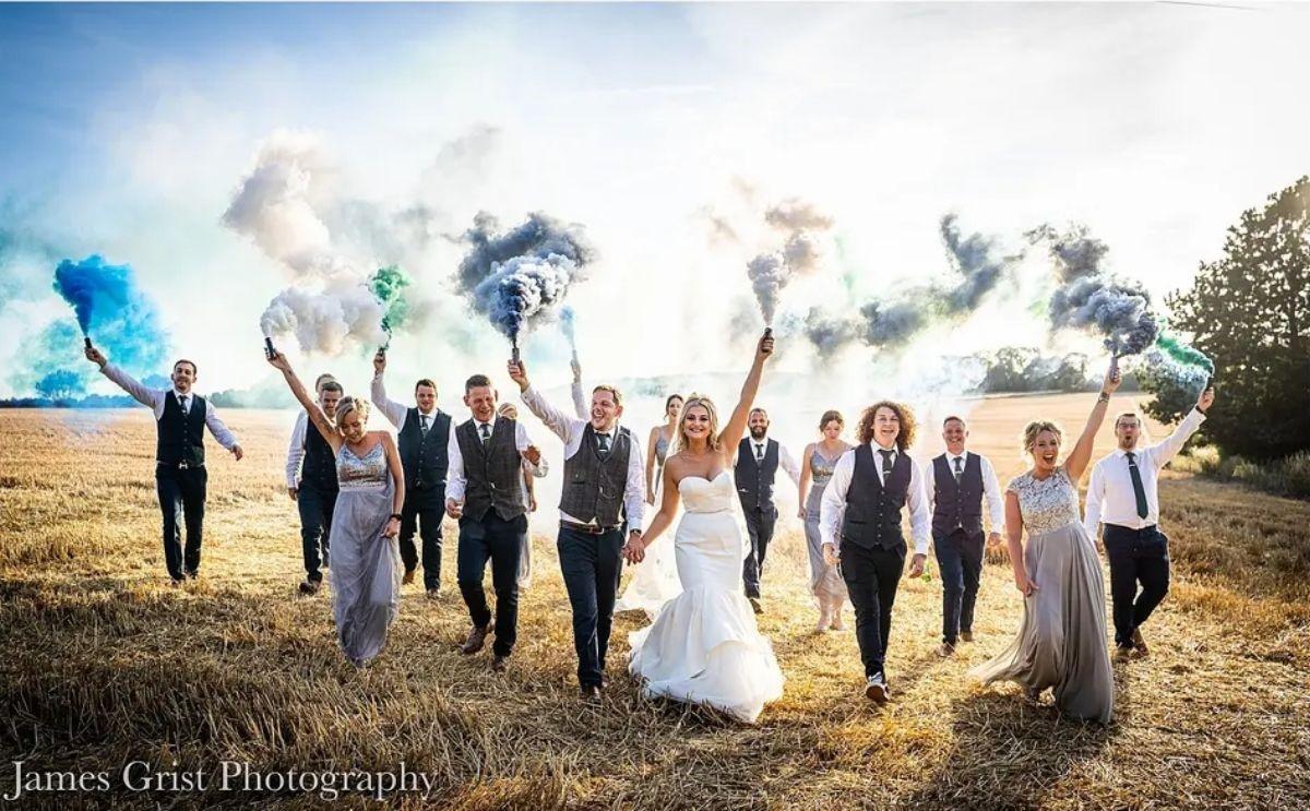 a bride, groom and large wedding party all walking towards the camera holding smoke cannons