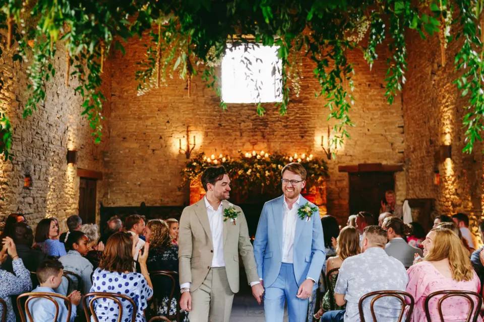 A groom in a beige wedding suit holding hands with his husband who is wearing a light blue wedding suit as they walk down the aisle together
