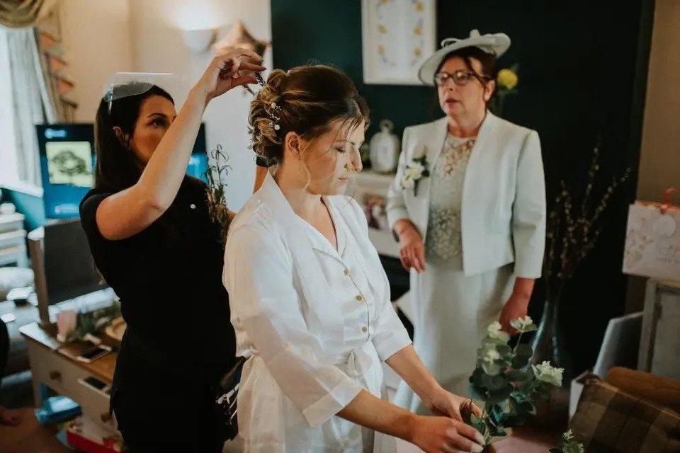 A standing bride arranges flowers while a hair stylist inserts an accessory into her hair