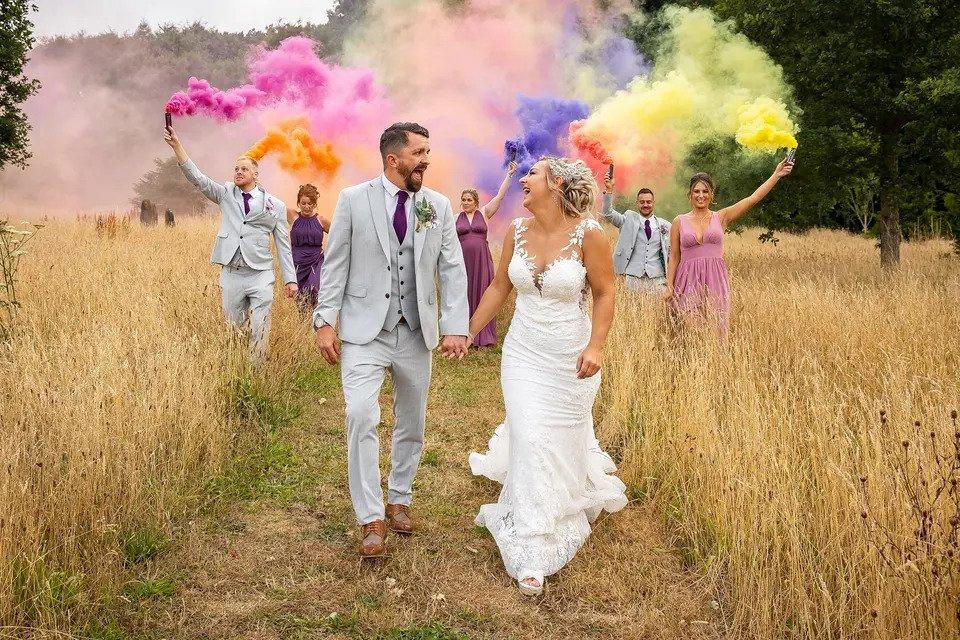 bride and groom walk hand in hand through a wheat field with the wedding party behind spraying coloured powder dye