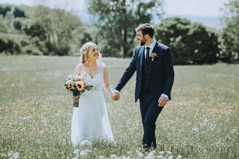 Bride and groom smiling and holding hands as they walk through the grounds of a wedding venue. The bride carries a bouquet of sunflowers and the groom has a sunflower buttonhole