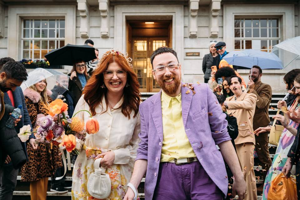 A bride wears a cream blouse and floral skirt with her husband in a purple suit as they walk down the stairs of a town hall after their wedding