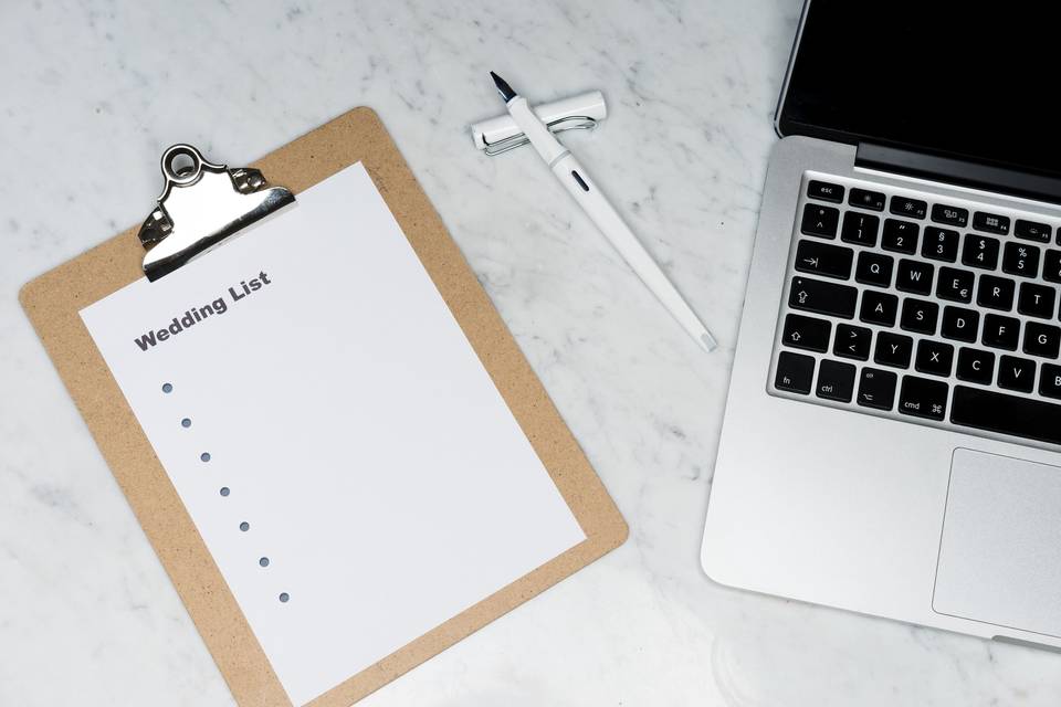 A brown clipboard with a wedding list next to a silver laptop couples can use to budget for additional wedding costs