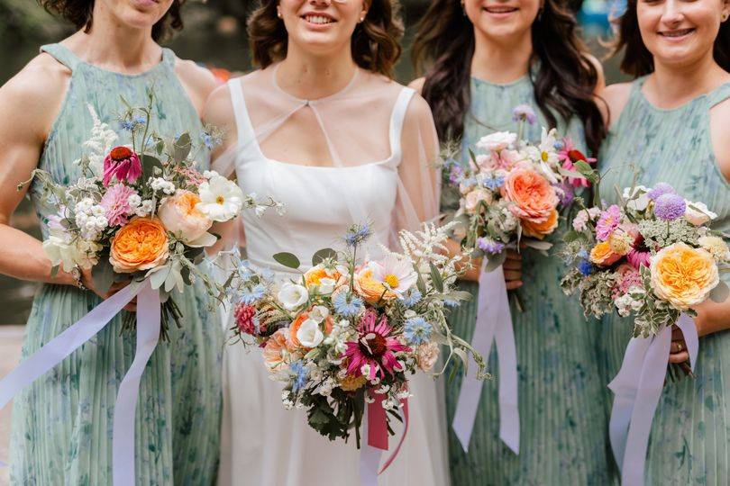 A bride surrounded by three bridesmaids dressed in sage green, each holding a summer bouquet of apricot, pink and cream wedding flowers, all smiling at the camera.