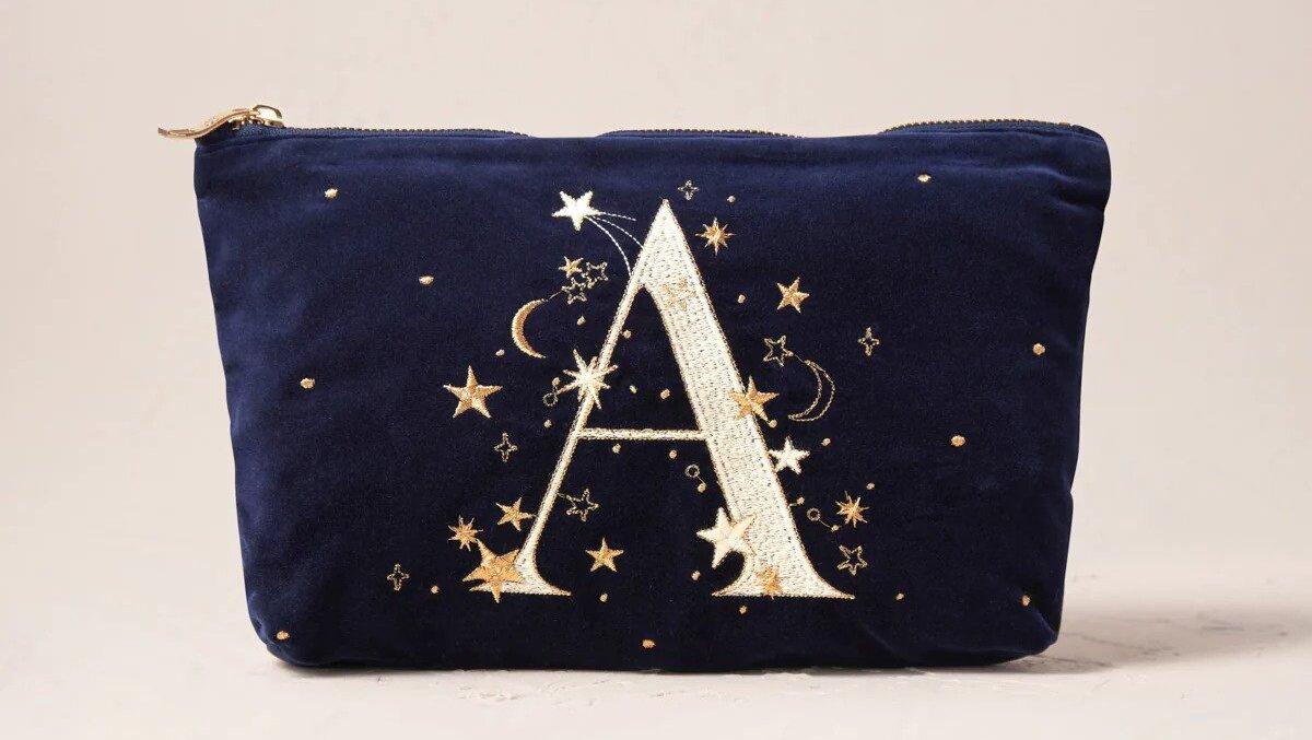 A velvet makeup bag with an 'A' embroidered on to a navy blue background, decorated with embroidered stars and moons