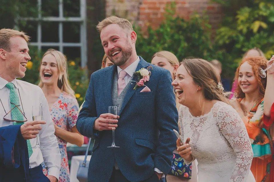 bride and groom stand outdoors laughing with guests and drinking champagne