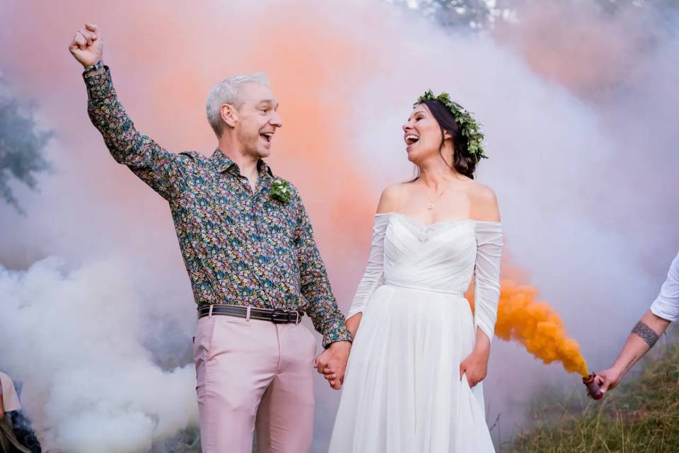 a bride and groom holding hands - the bride is wearing a wedding dress and flower crown and the groom is wearing a dark floral shirt and beige chino trousers - there is pastel pink smoke bombs behind them