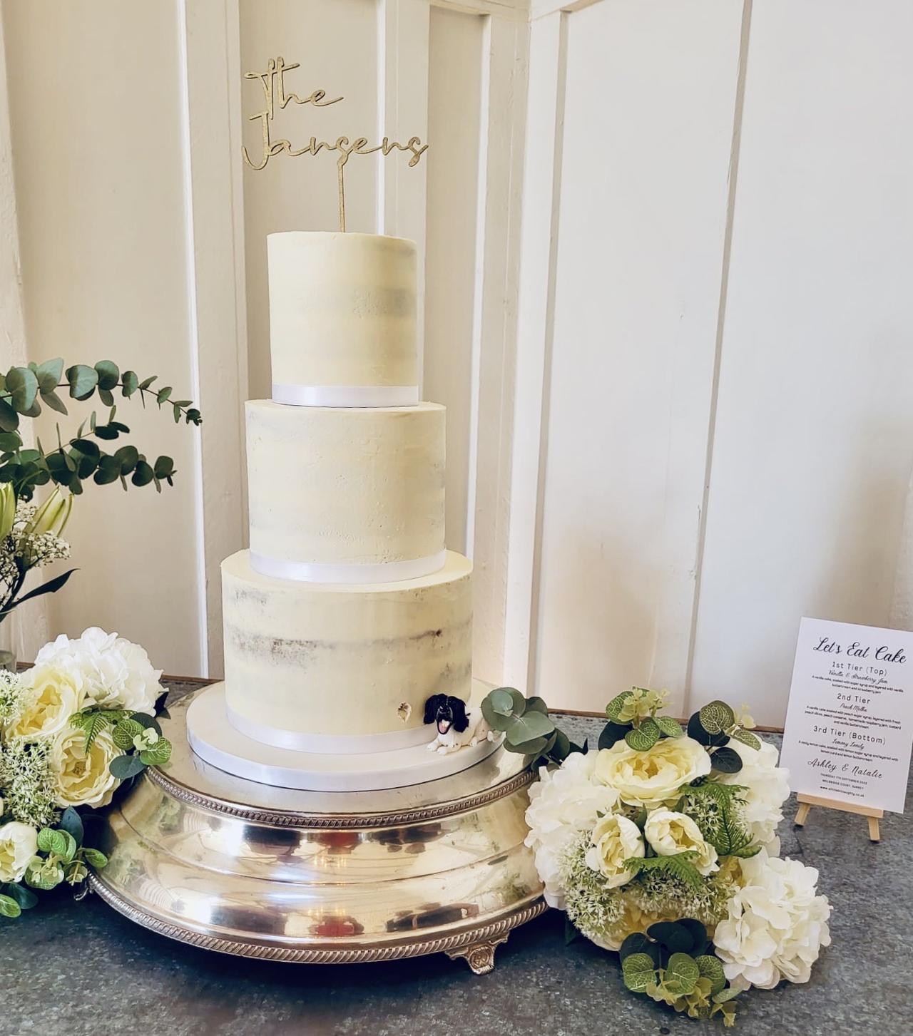 A white buttercream semi naked tiered cake on a silver cake stand.