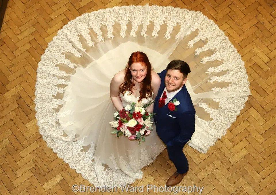 Photo of a bride and groom taken from above, as they look up at the camera. the bride's dress is fanned out around her highlighting the lace trim