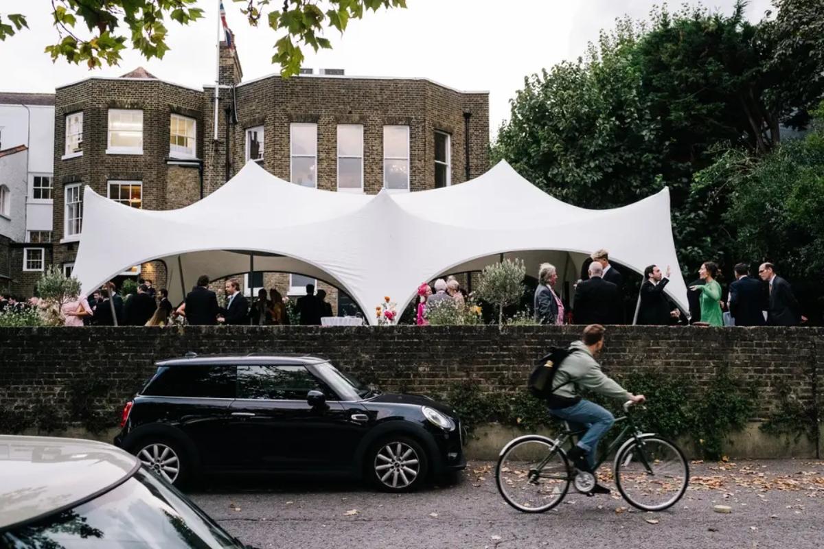  a wall with a car parked near it and a bicycle riding past - behind the wall is a marquee and a small london wedding venue with guests enjoying drinks outside