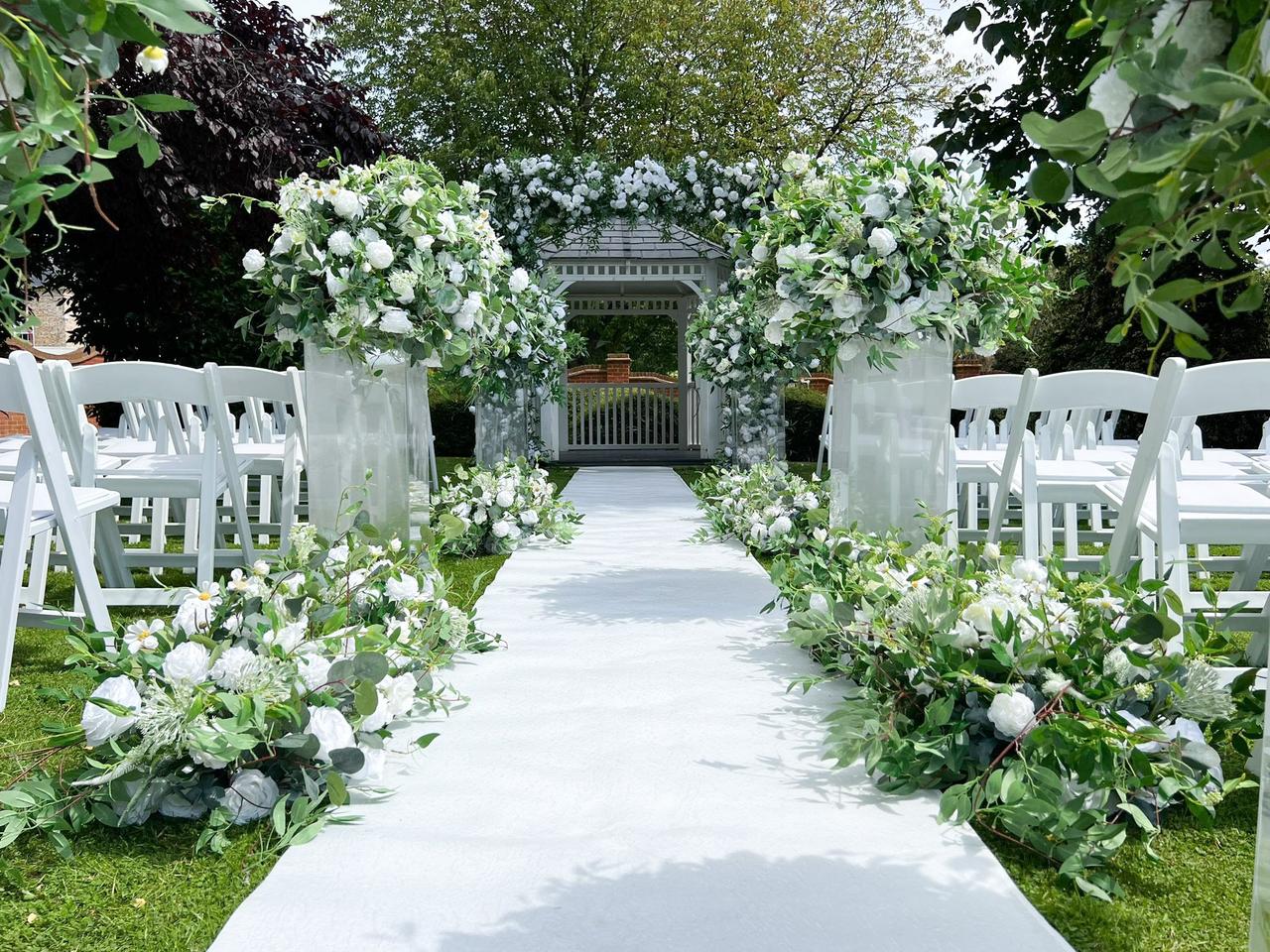 Outdoor wedding ceremony decorated with white chairs, a white aisle runner, white flowers and greenery, leading up to a wooden pagoda