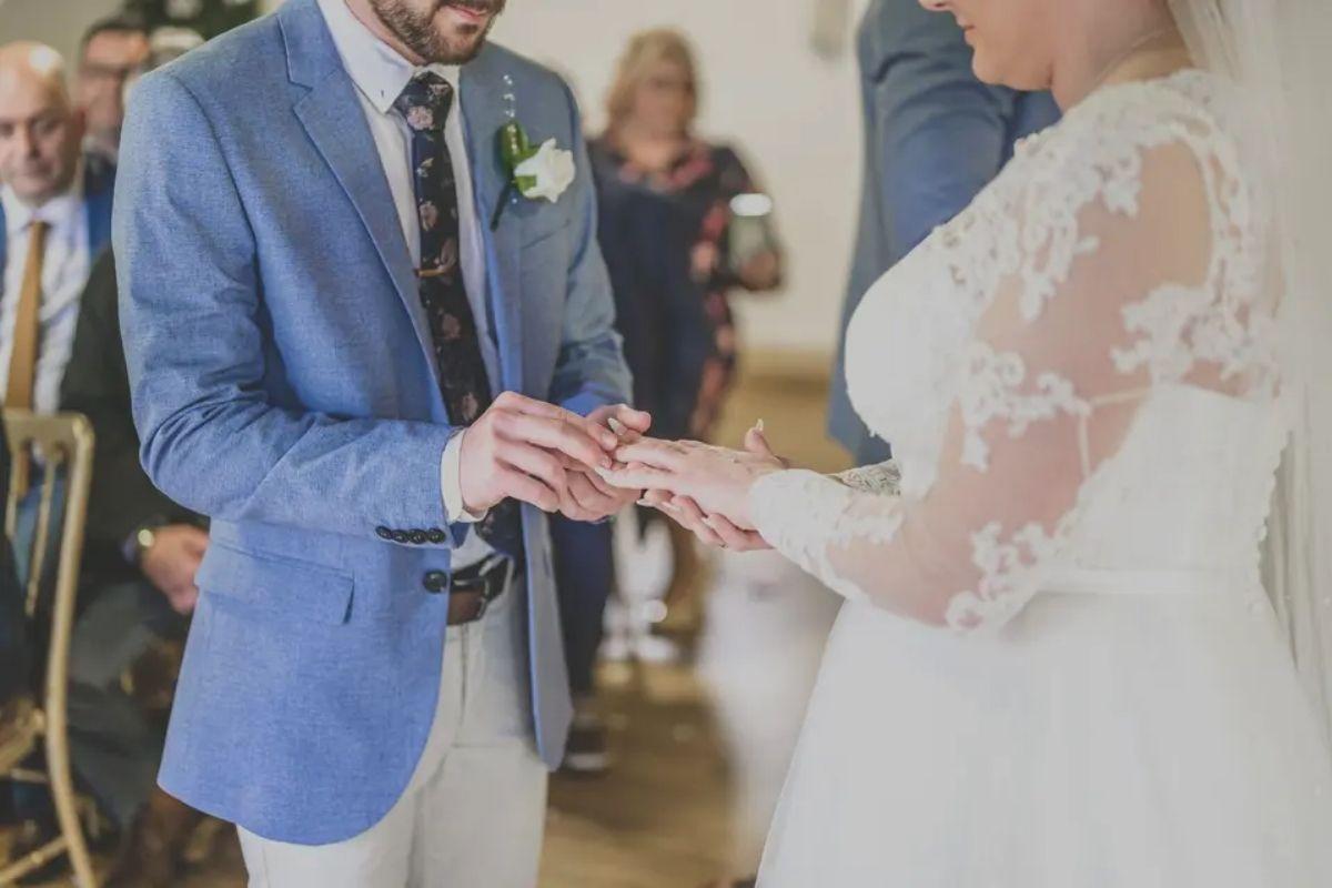 a shot showcasing a groom putting a wedding ring on his partner's finger as they both stand at the altar