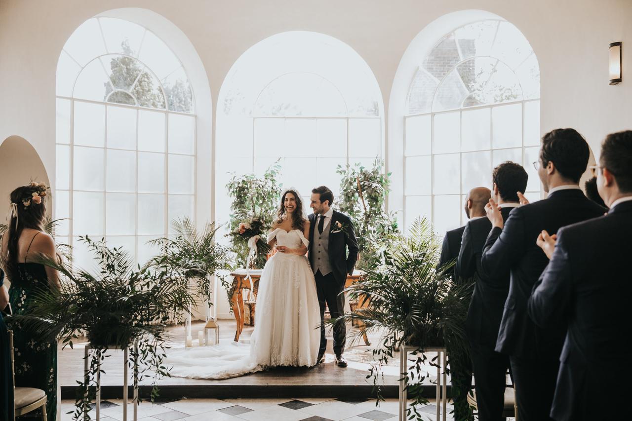 Bride and groom standing in front of their friends at a white alter in front of three tall French windows