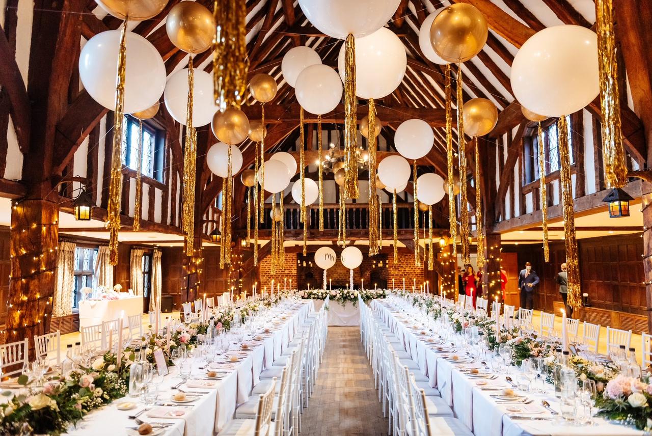 Interior shot of the barn at Great Fosters adorned with ivory and gold oversized balloons and two long banqueting tables
