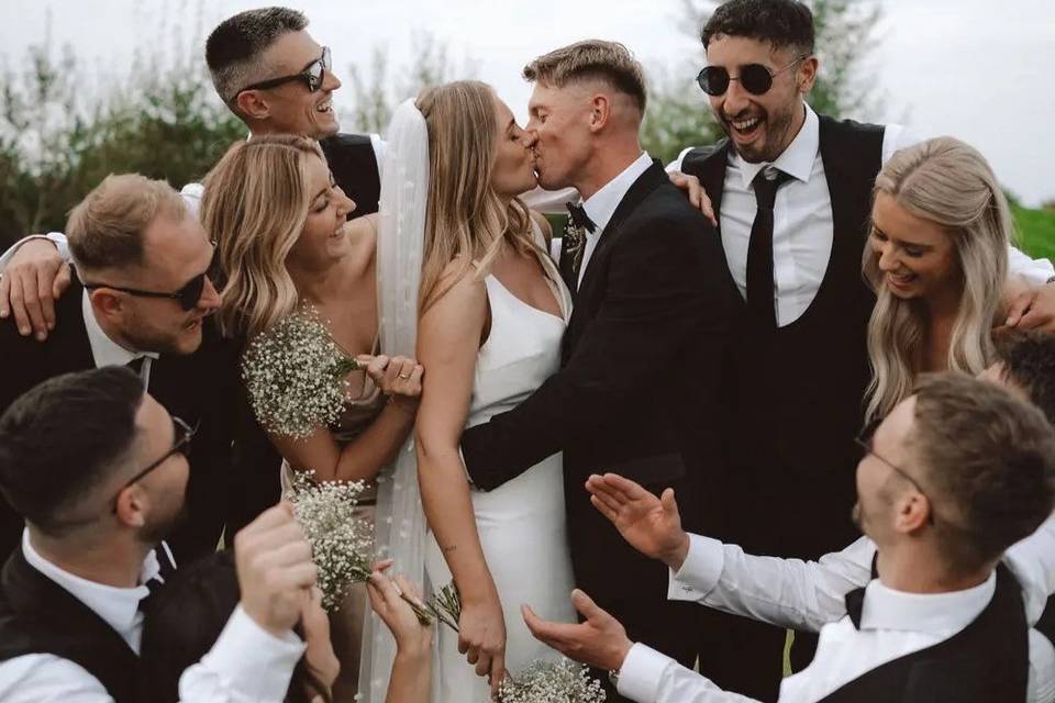 A newly married bride and groom kissing after their wedding ceremony as their wedding party surrounds them smiling and laughing
