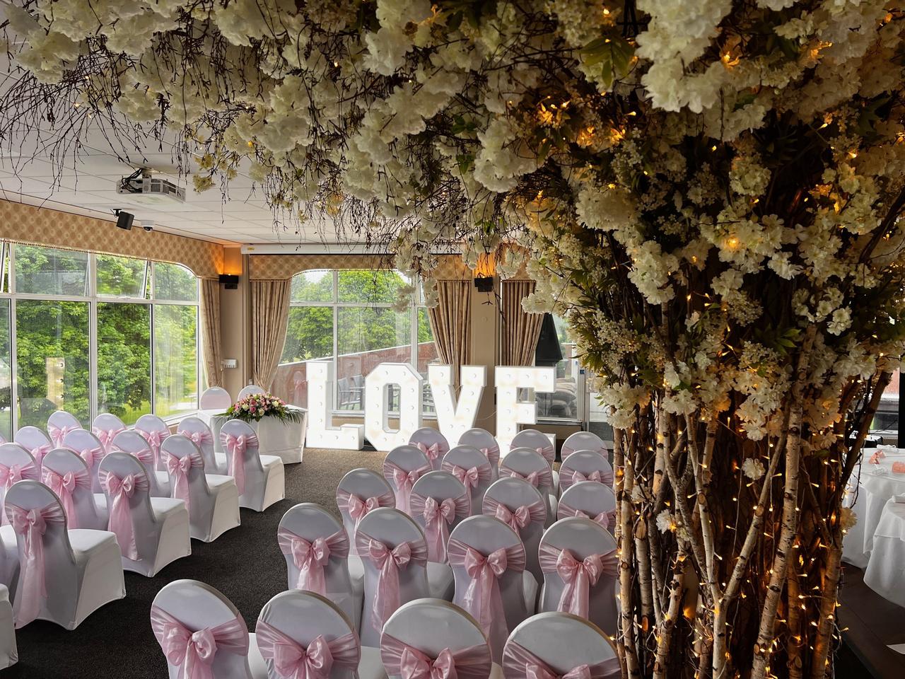 Interior of a room set up for a wedding ceremony with chair covers and pink bows and a light-up LOVE sign.