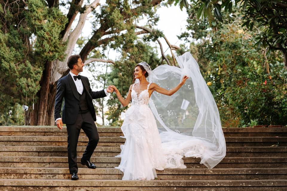 A bride and groom hold hands and smile at each other while descending stone stairs and the bride throws her veil out behind her. 