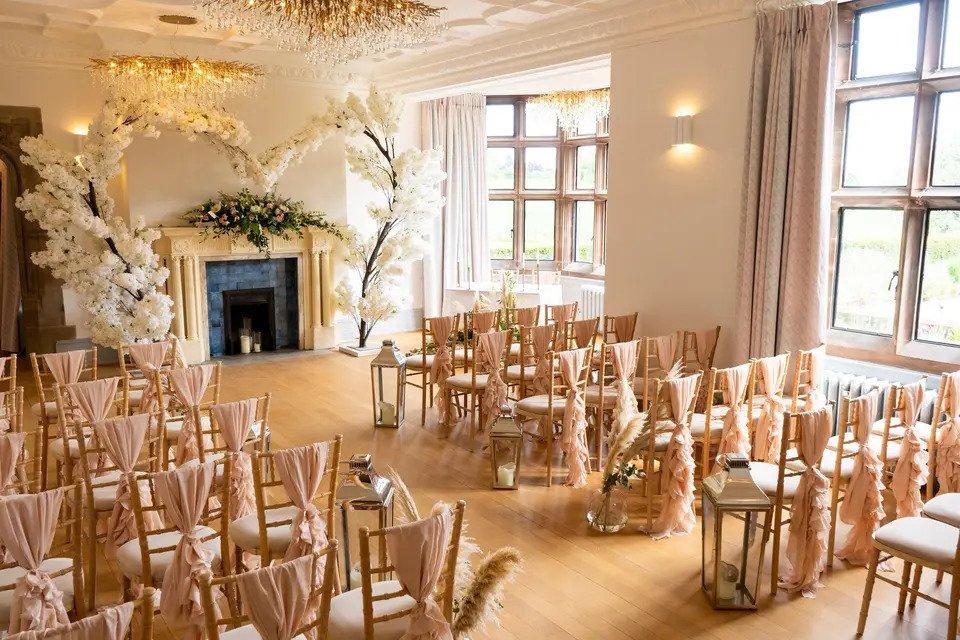 Room set for wedding ceremony with Chiavari chairs, chair sashes, pampas grass, fireplace and floral arch