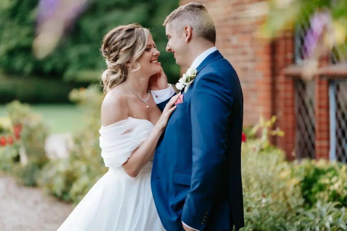 a bride wears an off the shoulder white wedding dress as she smiles and faces her groom outside their wedding venue