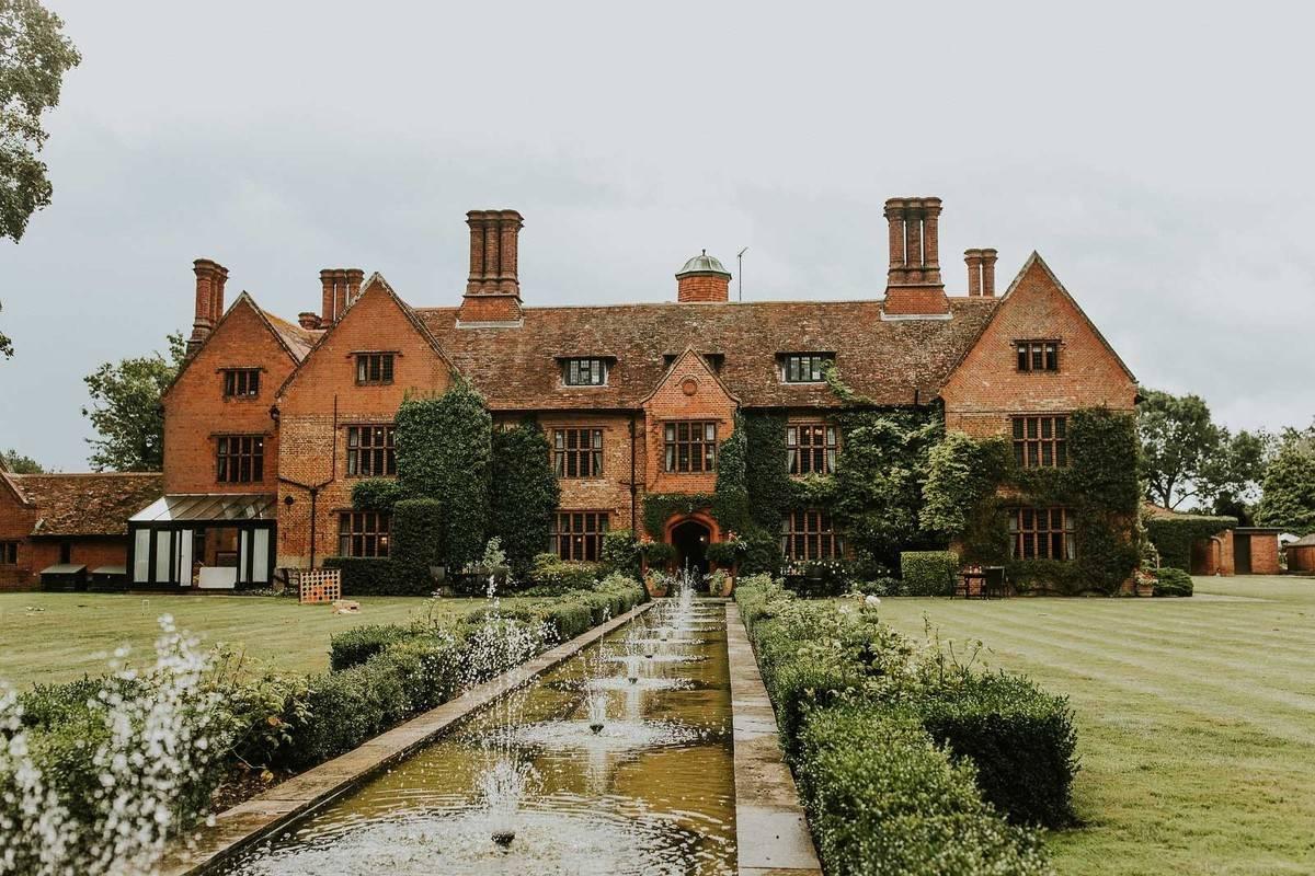 A front view of brick built Suffolk wedding venue Woodhall Manor with a long rectangular pond at the front 