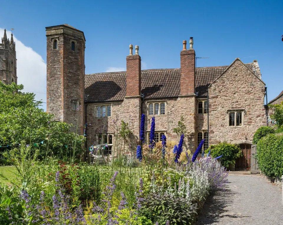 Grey stone farmhouse venue with twin chimneys and a verdant lawn full of flowers beside a traditional church