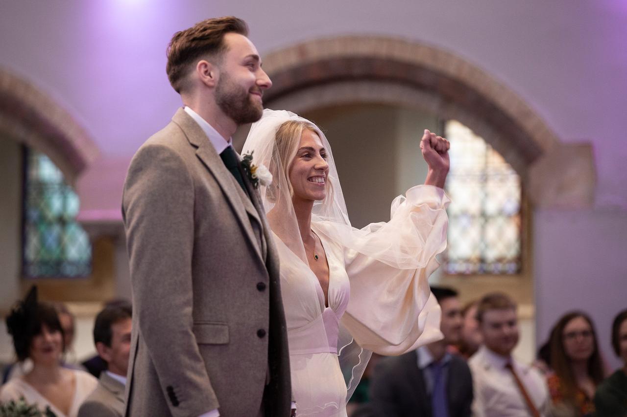 A bride and groom look on as their Christian wedding ceremony takes place. The bride is smiling with her hands in the air for worship.