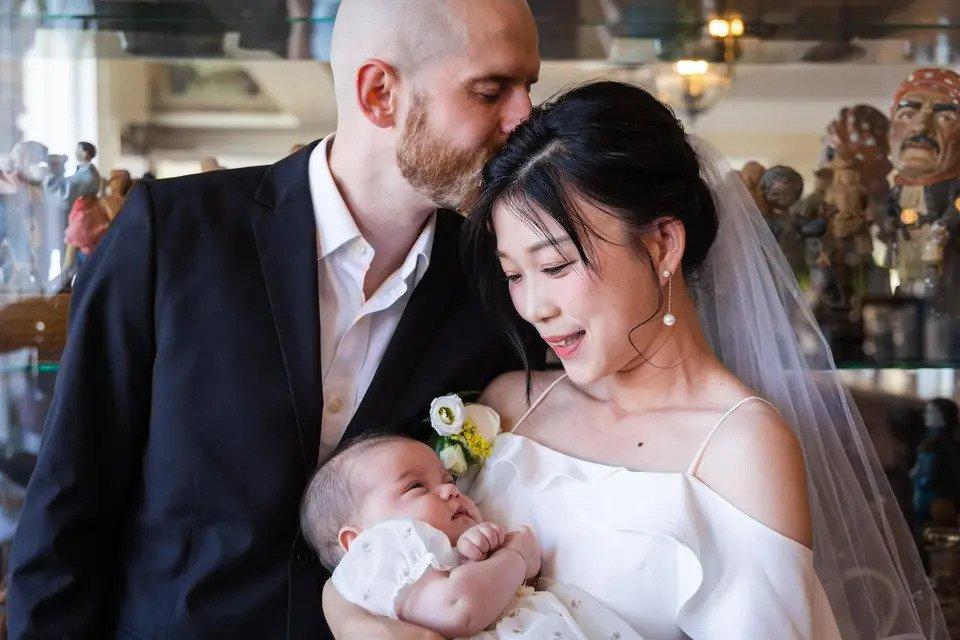 Bride and groom hold their baby while groom kisses the brides head