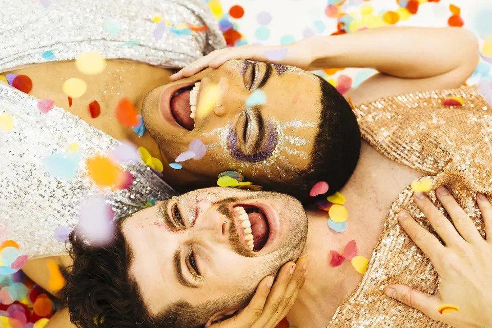 A queer couple lie down surrounded by multi-coloured confetti smiling