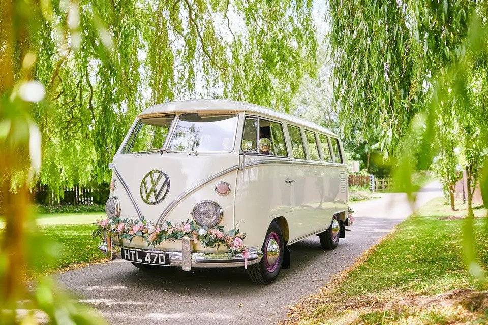 A cream VW camper van decorated with pink flowers parked outside surrounded by grass and trees